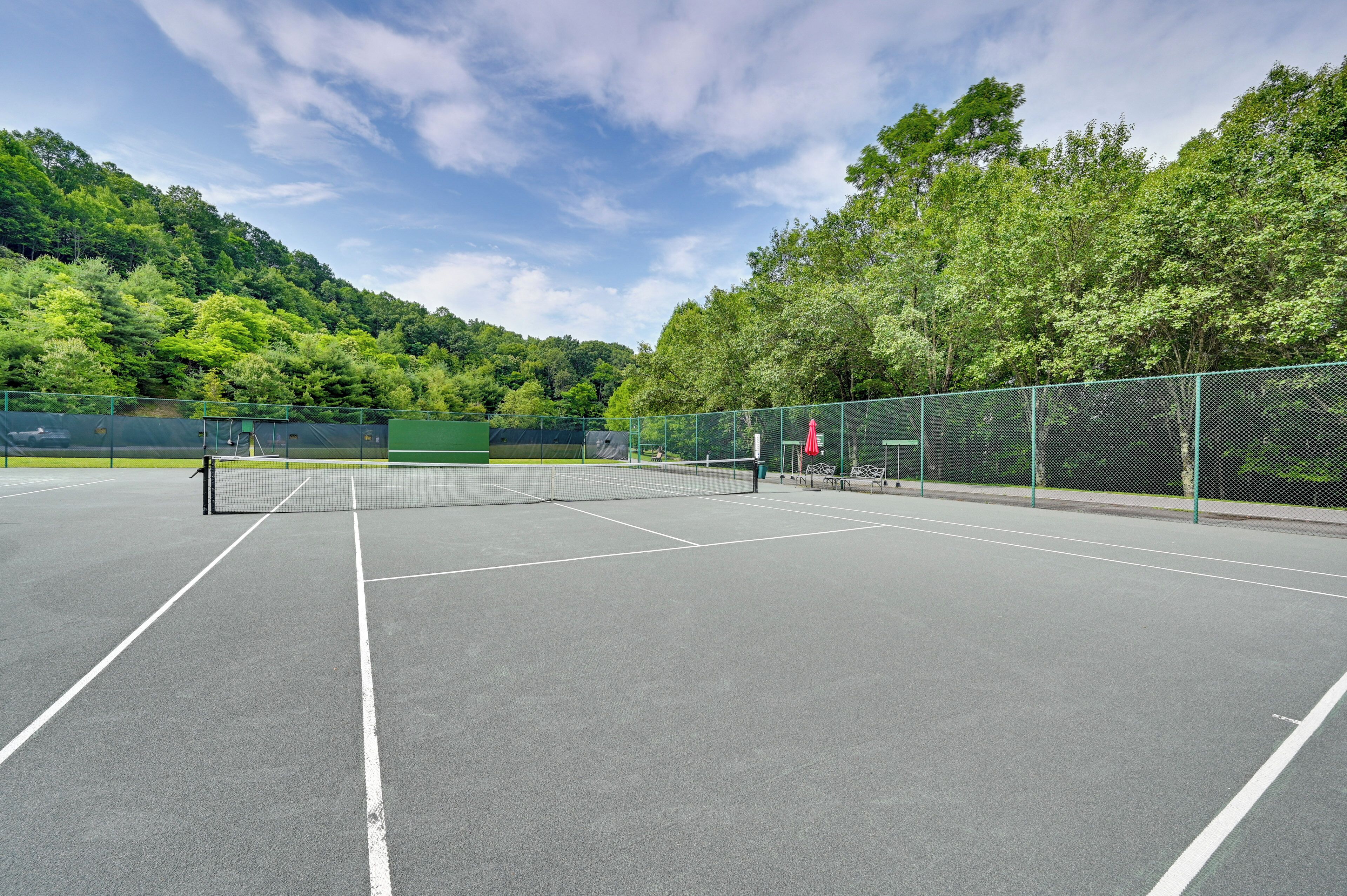 An outdoor tennis court with a net, surrounded by green fencing and trees.