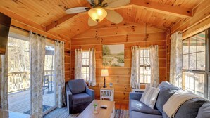 A cozy cabin interior with wooden walls, a ceiling fan, a sofa, a chair, a small table, and a window with curtains. - Steps to Virginia Creeper Trail: Creekside Retreat (Damascus)
