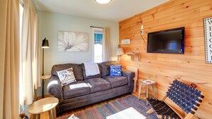 A cozy living room with a brown leather sofa, a small wooden table, a blue woven bench, and a flat-screen TV mounted on a wooden wall.