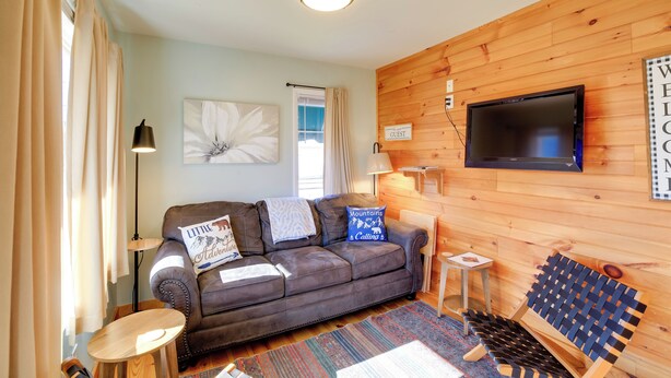 A cozy living room with a brown leather sofa, a small wooden table, a blue woven bench, and a flat-screen TV mounted on a wooden wall. - Trail Town Usa! 2BD Cabin on Laurel Creek w/ Deck (Damascus)