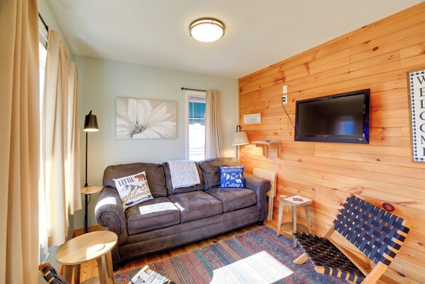 A cozy living room with a brown leather sofa, a small wooden table, a blue woven bench, and a flat-screen TV mounted on a wooden wall. - Trail Town Usa! 2BD Cabin on Laurel Creek w/ Deck (Damascus)