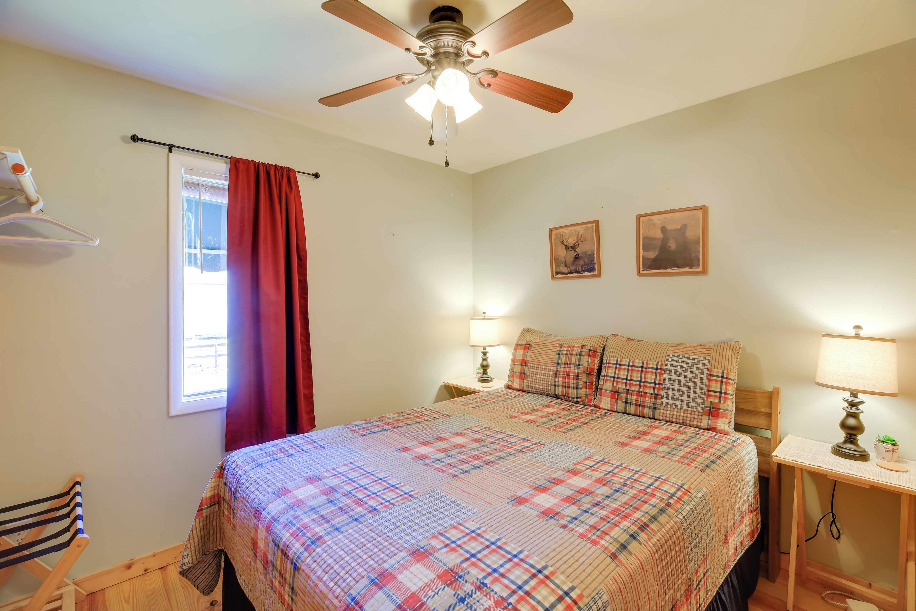 A bedroom with a bed, a ceiling fan, a window with red curtains, two bedside lamps, and a small table with a plant.