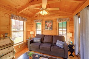 A cozy cabin interior with a wooden ceiling, a ceiling fan, a brown sofa with pillows, a side table with a lamp, and a window with plaid curtains. - Pet-friendly Damascus Cabin w/ Deck & Mtn Views! (Damascus)