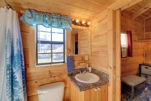Un baño de cabaña de madera con un inodoro blanco, un lavabo con encimera de granito, un espejo y una ventana con cortina.
