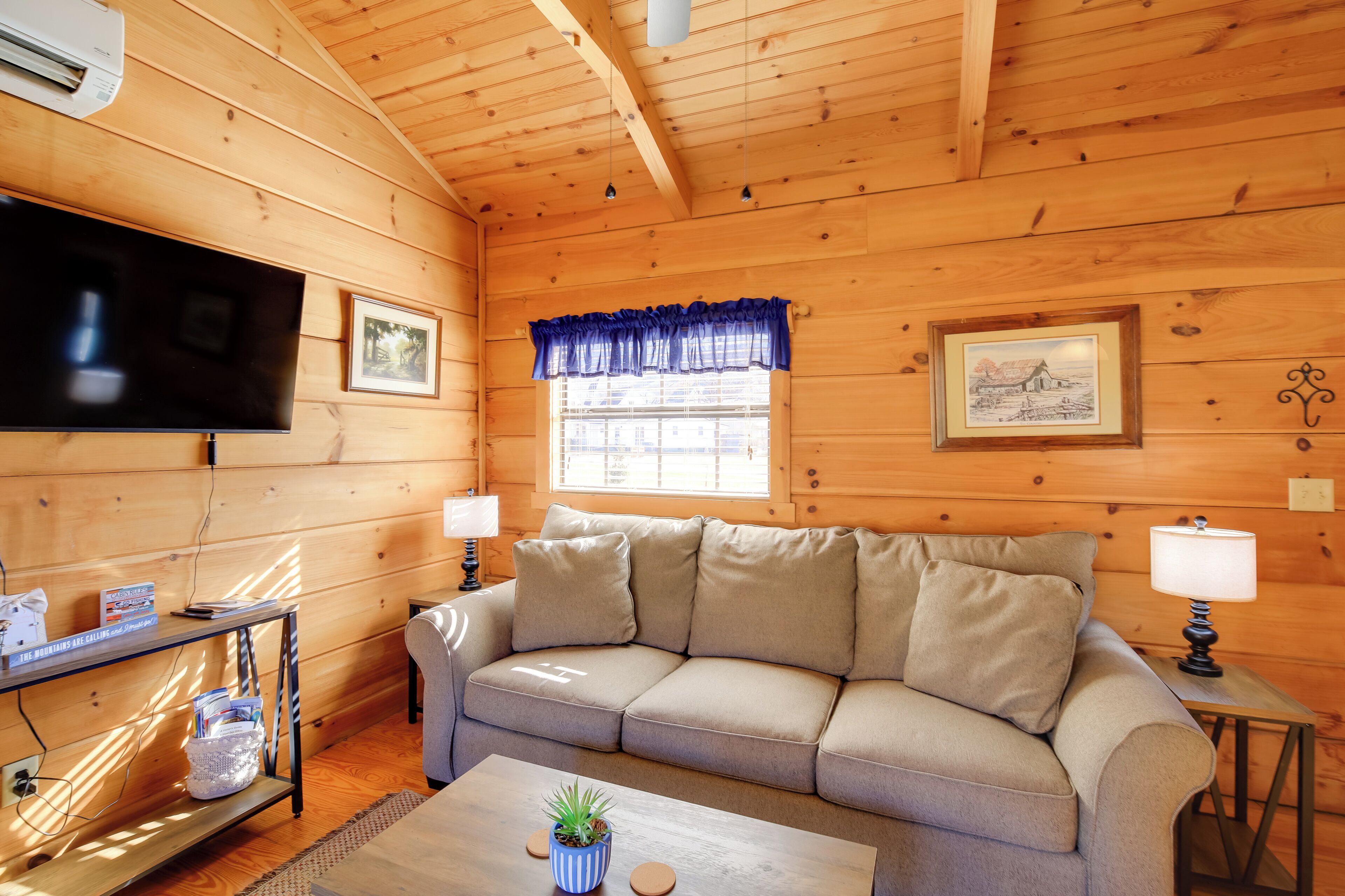 A cozy wooden cabin interior with a sofa, a flat-screen TV, a table with a plant, and a window with a blue valance.