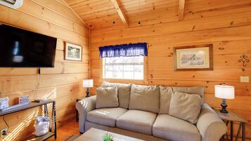 A cozy wooden cabin interior with a sofa, a flat-screen TV, a table with a plant, and a window with a blue valance.
