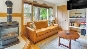 A cozy living room with a fireplace, a leather sofa, a wooden coffee table, and a bookshelf with books and a basket. - Ludlow Cabin on Lake Pauline w/ Outdoor Fire Pit (Ludlow)