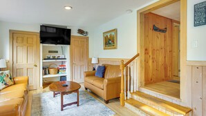A cozy living room with a brown leather sofa, a wooden coffee table, and a bookshelf. A staircase leads to an upper level. - Ludlow Cabin on Lake Pauline w/ Outdoor Fire Pit (Ludlow)