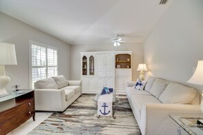 Ein helles Wohnzimmer mit einem weißen Sofa, einem Glastisch und einem Holzkasten.