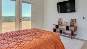 Un dormitorio con cama, televisor montado en la pared, estante de madera con objetos decorativos y vista a un viñedo a través de puertas correderas.