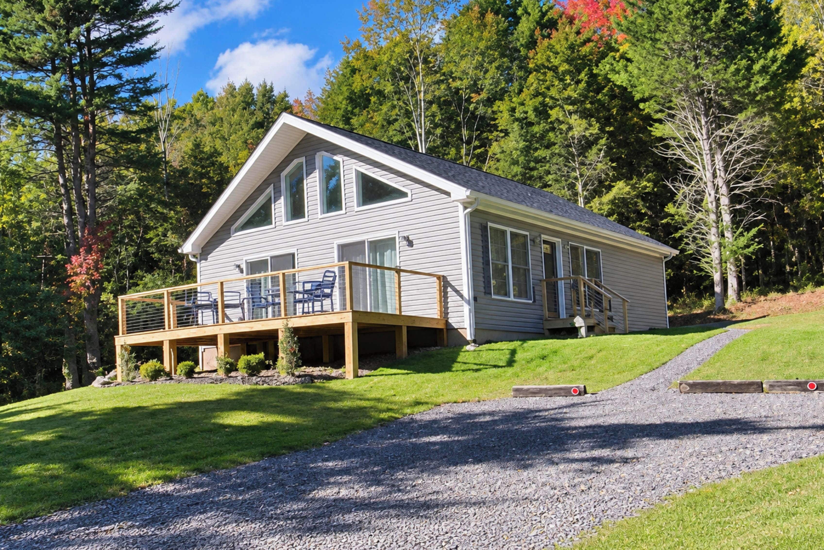 A two-story house with a deck and a covered porch, surrounded by trees and a grassy hill.