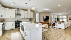 A modern kitchen with white cabinets, a central island, and stainless steel appliances. There is a dining area with a wooden table and chairs, and a living room with a white sofa and a TV. - Walk to Keansburg Beach! Family Home w/ Pool (Keansburg)
