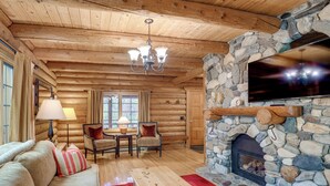 A log cabin interior with a stone fireplace, wooden ceiling beams, a television mounted on the wall, and a cozy seating area with a sofa and chairs. - Along Big Wood River: Hailey Family Cabin w/ Yard (Hailey)