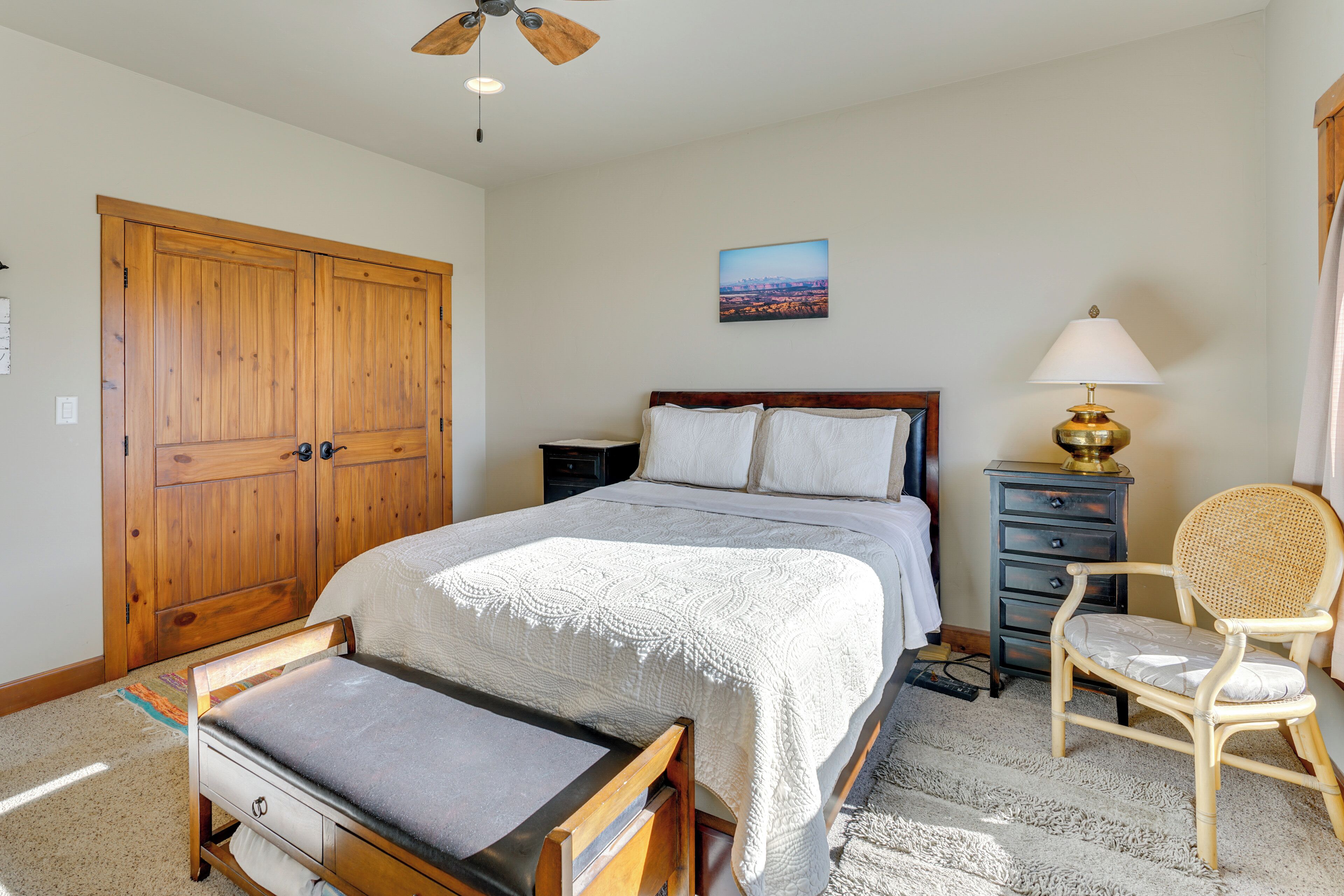 A bedroom with a large wooden wardrobe, a bed with a white bedspread, a nightstand with a lamp, a wicker chair, and a cityscape painting on the wall.