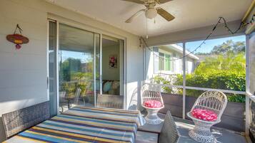 A covered outdoor balcony with a bed, two wicker chairs, and a ceiling fan.