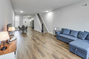 A modern living room with a wooden floor, a grey sofa, a dining area with a table and chairs, and a staircase leading to another level. - Pet-friendly 3BR Rowhouse: Walk to Patterson Park! (Baltimore)