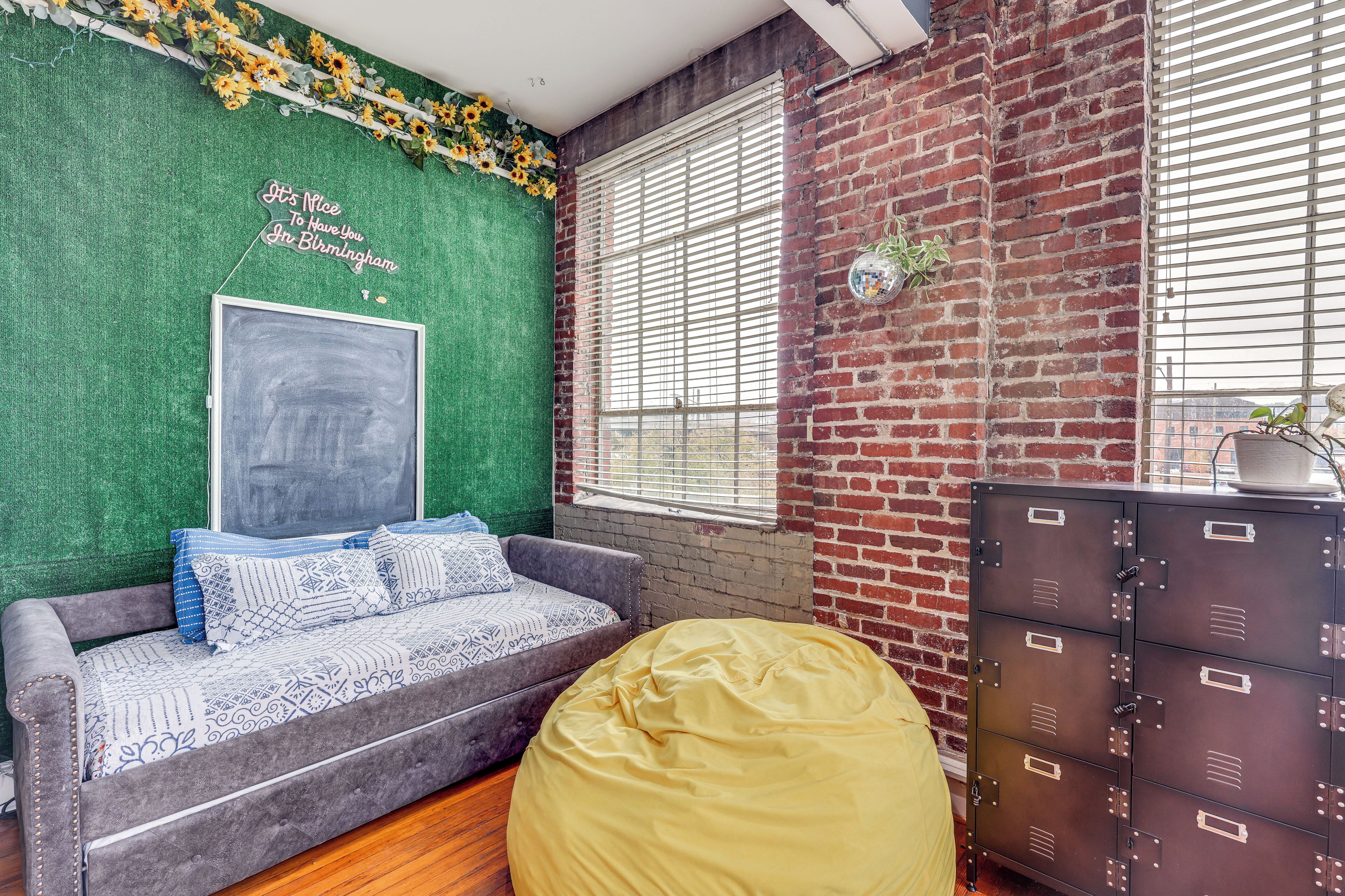 A room with a green wall, a grey sofa with patterned cushions, a yellow bean bag, a metal locker, and a window with blinds.