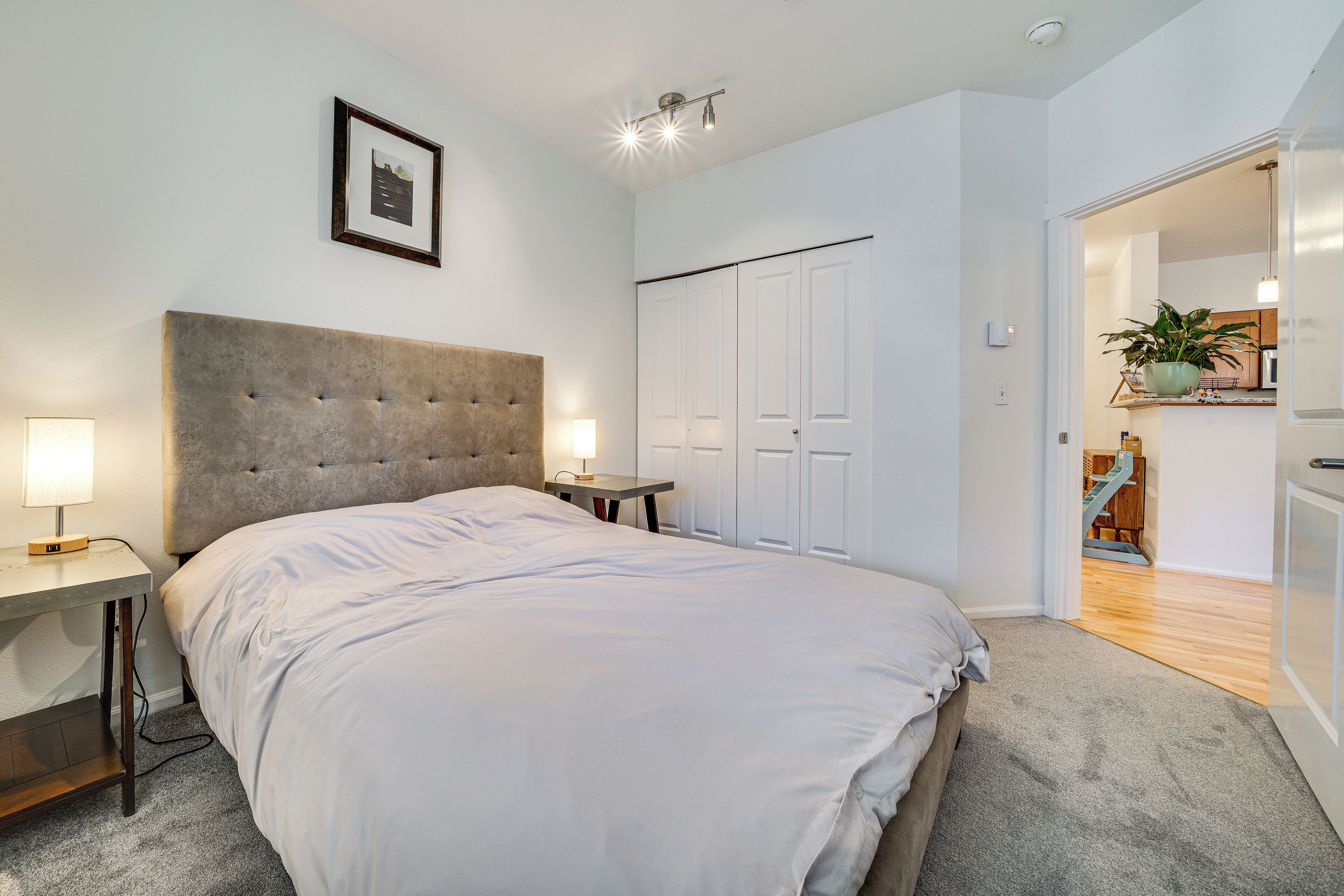 A bedroom with a large bed, a bedside table with a lamp, a framed picture on the wall, and a closet with a door.