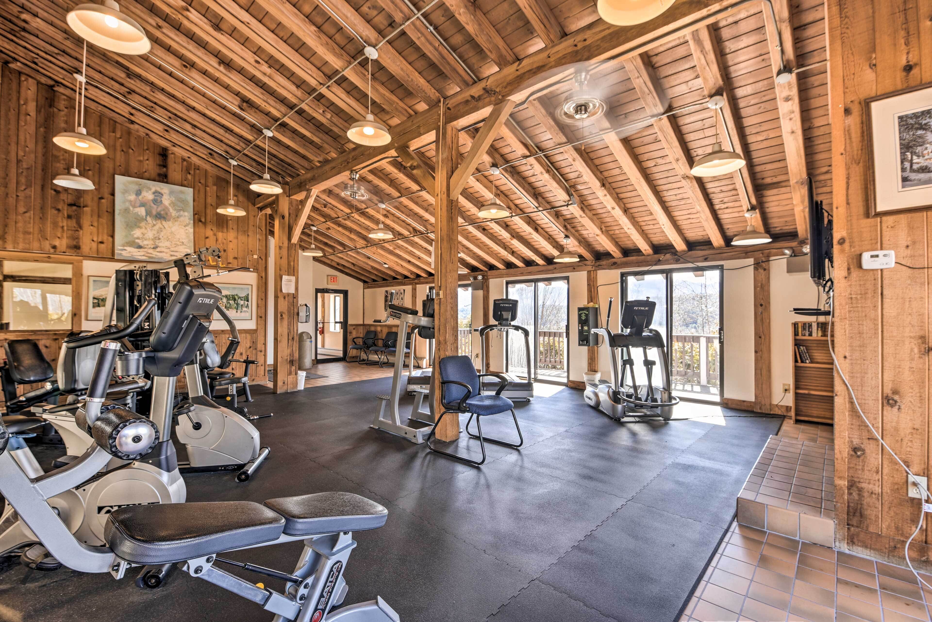 Un gimnasio con diversos equipos de ejercicio, vigas de madera en el techo y amplios ventanales que dejan entrar luz natural.