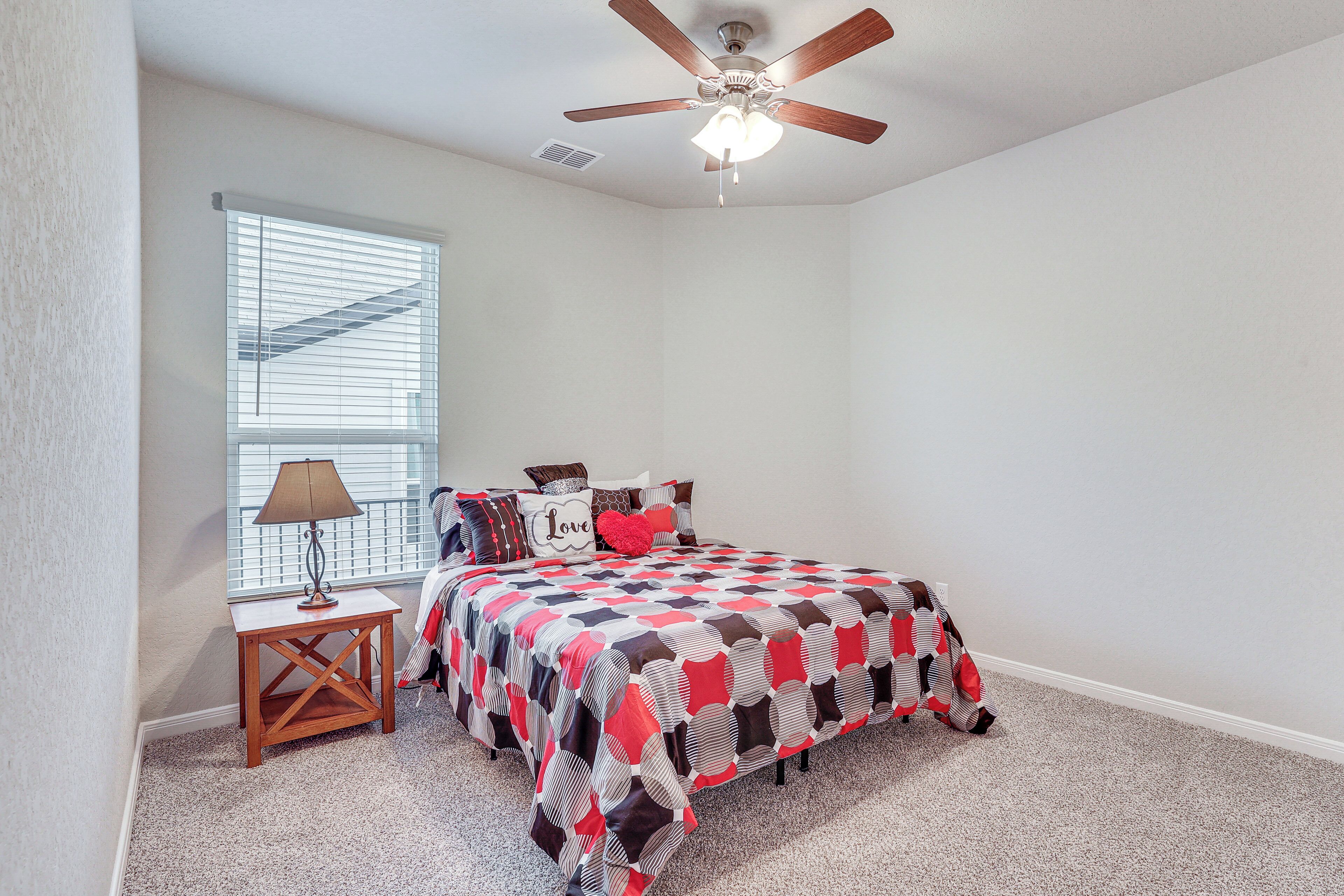 A bedroom with a bed, a ceiling fan, a window with blinds, and a nightstand with a lamp.