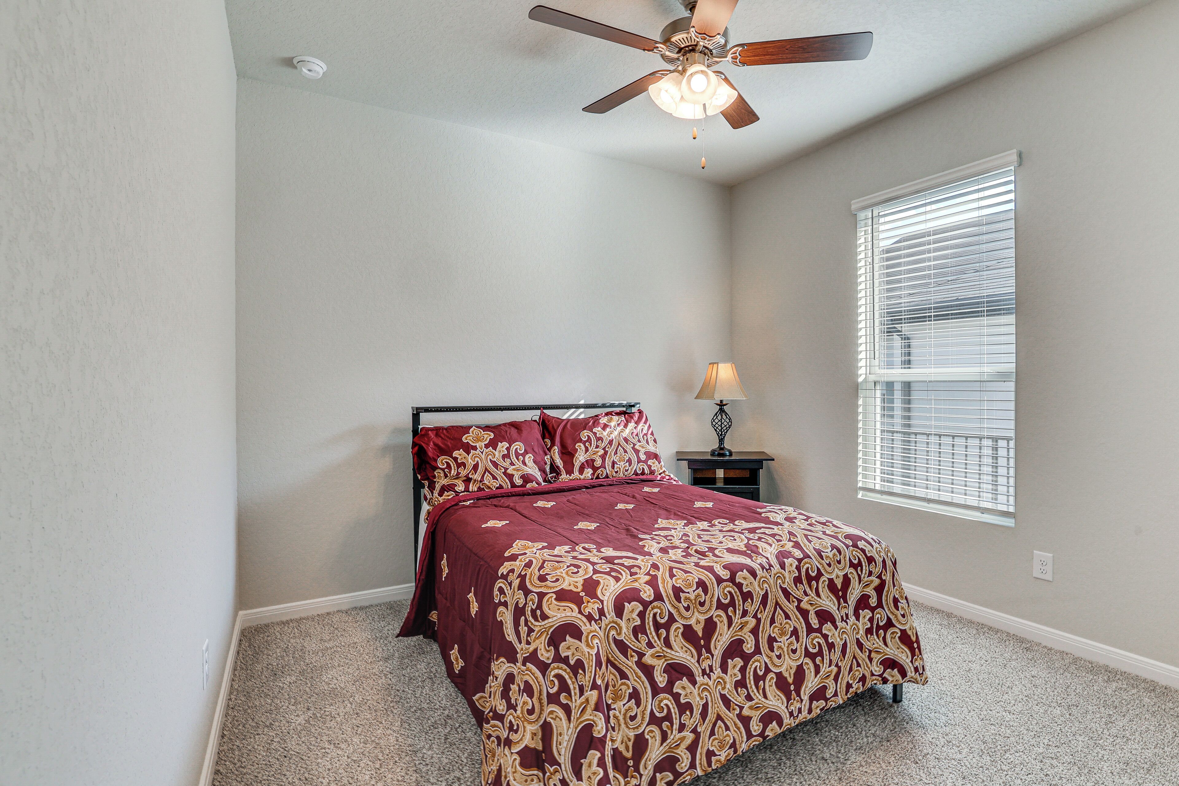 A bedroom with a bed, a ceiling fan, a window with blinds, and a nightstand with a lamp.