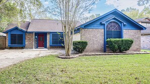 A two-story house with a blue exterior, a red door, and a small front yard with a tree and bushes. - Extended Stay in Sugar Land! Family Home w/ Yard (Sugar Land)