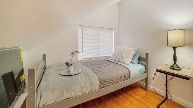 Ein kleines Zimmer mit einem Einzelbett, schrägen Decken, einem Fenster mit Jalousien, einem Nachttisch mit Lampe und einer kleinen Pflanze auf dem Bett.