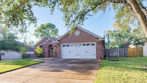 A two-story brick house with a white garage door, a front yard with grass, and a tree providing shade. - Gateway to Aggieland! College Station Family Home (College Station)