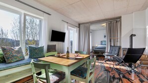 Une pièce avec une table en bois, des chaises vertes, un canapé avec des coussins à motifs, un téléviseur et une grande fenêtre avec des rideaux.