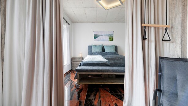 Une chambre à coucher comprenant un lit, des rideaux, une tête de lit, une table de chevet et un luminaire fixé au mur.