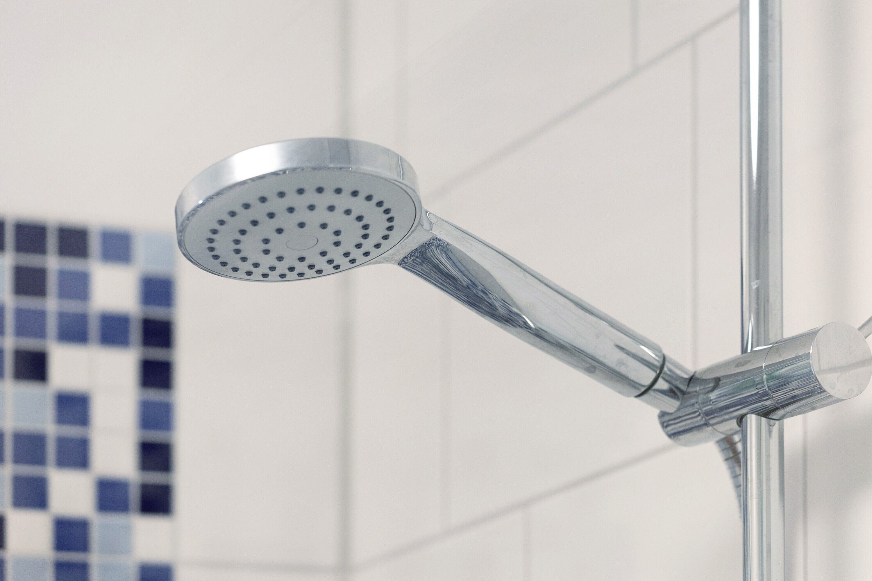 Un cabezal de ducha cromado unido a un tubo vertical en un baño con paredes de azulejos blancos.