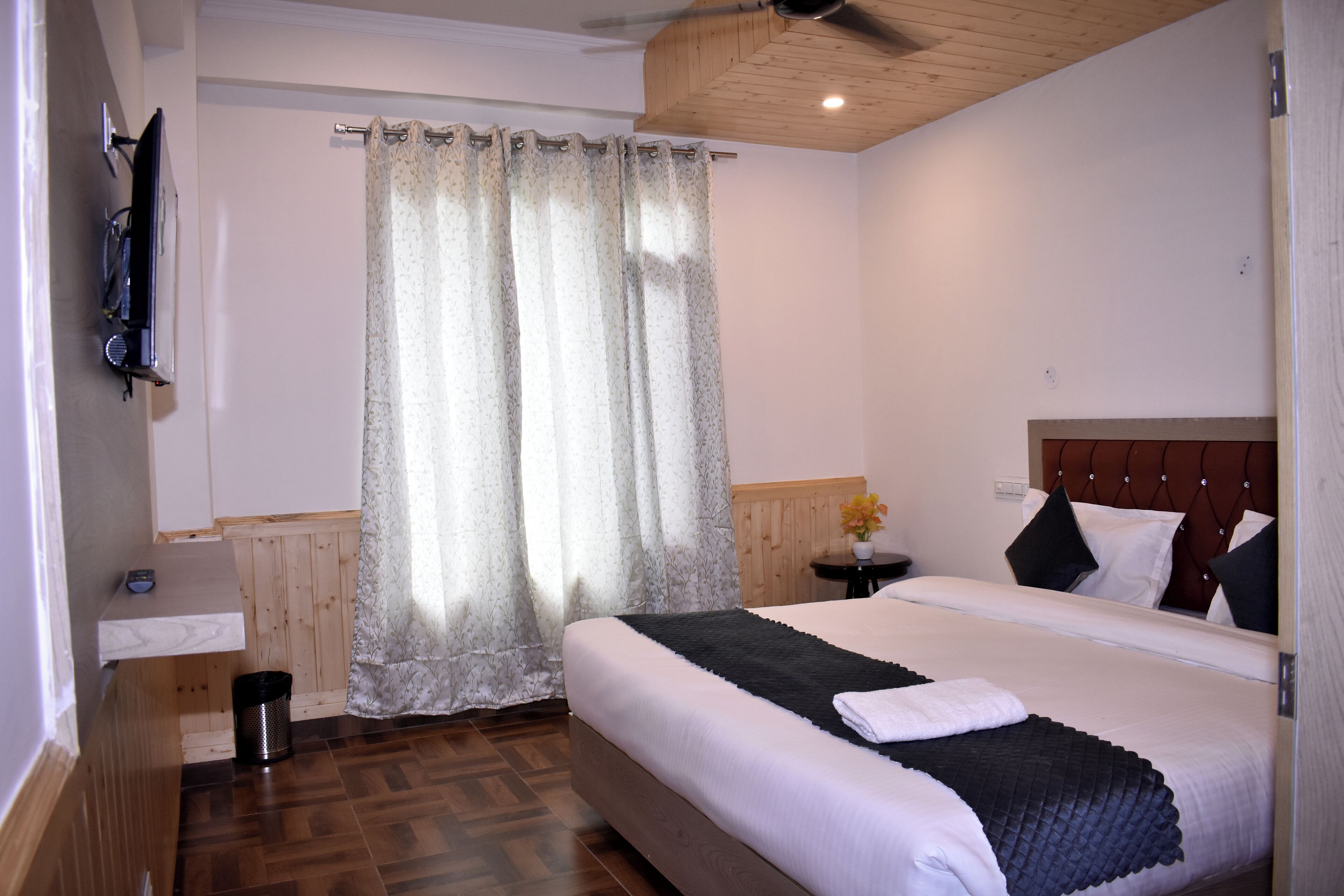 Un dormitorio con suelo de madera, una cama con cabecera oscura, una mesita de noche con lámpara y una ventana con cortina.