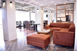 Ein modernes Wohnzimmer mit einem Ecksofa, einem Hocker und einem Glastisch. Im Hintergrund befindet sich ein Essbereich mit Stühlen und einem Tisch.