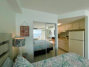 A bedroom with a bed, a bedside lamp, and a mirror. There is a kitchen area with cabinets and a refrigerator. A sign on the wall reads "Sandy toes & Salty kisses". - The Coastline | Oceanfront Studio | Salt Air (Daytona Beach)