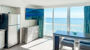 A modern kitchen with a refrigerator, a wooden cabinet, and a dining area with a table and chairs. A large window offers a view of the ocean. - The Atlantic | Direct Oceanfront | 10th Floor (Daytona Beach)
