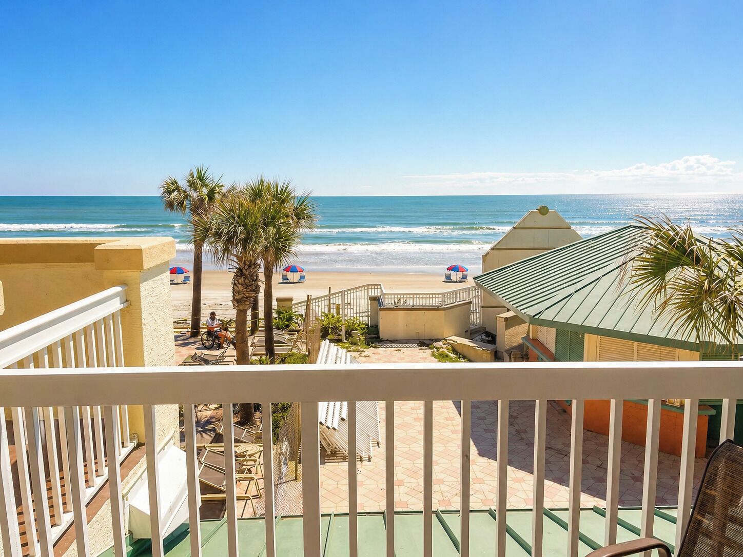 A balcony with a view of a beach, palm trees, and beach umbrellas.