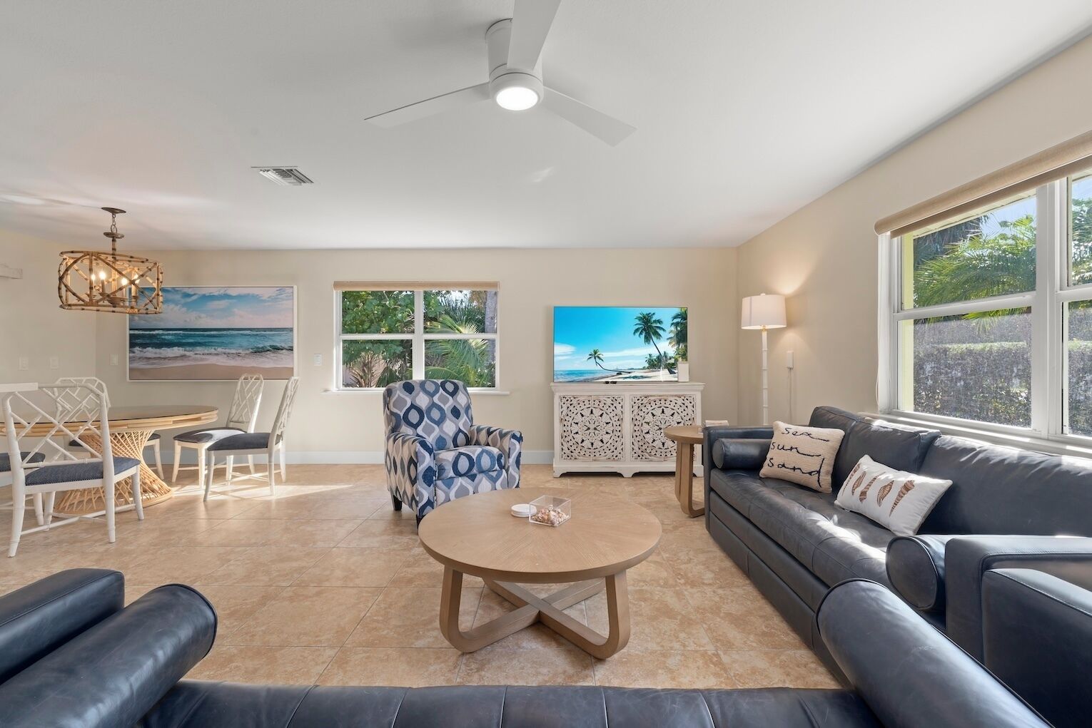 A spacious living room with a round coffee table, a sofa, a patterned armchair, and a dining area with a table and chairs. There are two windows with views of greenery and a beach.