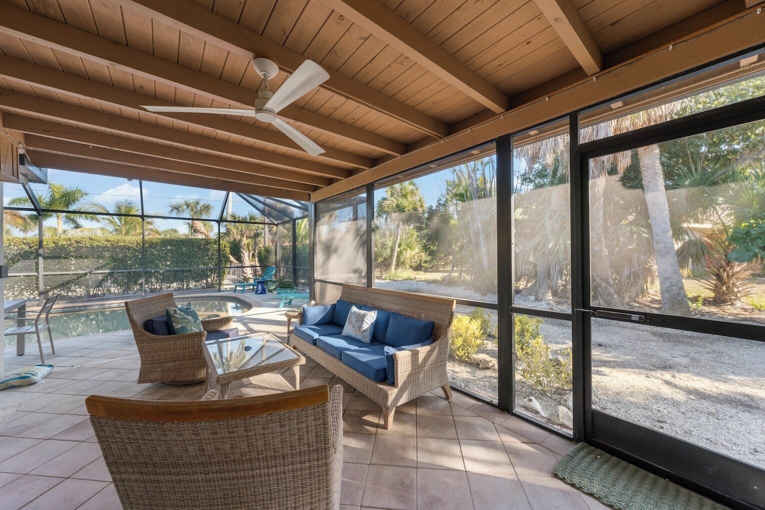 A covered outdoor seating area with wicker furniture, a glass table, and a ceiling fan.