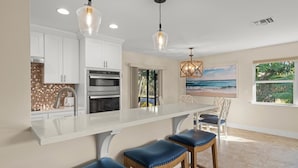 A modern kitchen with white cabinets, a large island with stools, and a view of the ocean through a window. - Tahitian Waters: Pet-Friendly Pool Home Just Steps to the Beach! (Sanibel)