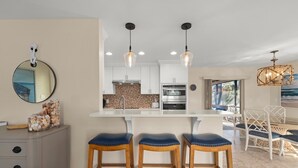 A modern kitchen with white cabinets, a built-in oven, and pendant lights. There are three wooden stools with blue cushions and a dining table with white chairs. A large mirror hangs on the wall, and a decorative wall piece is visible. - Tahitian Waters: Pet-Friendly Pool Home Just Steps to the Beach! (Sanibel)