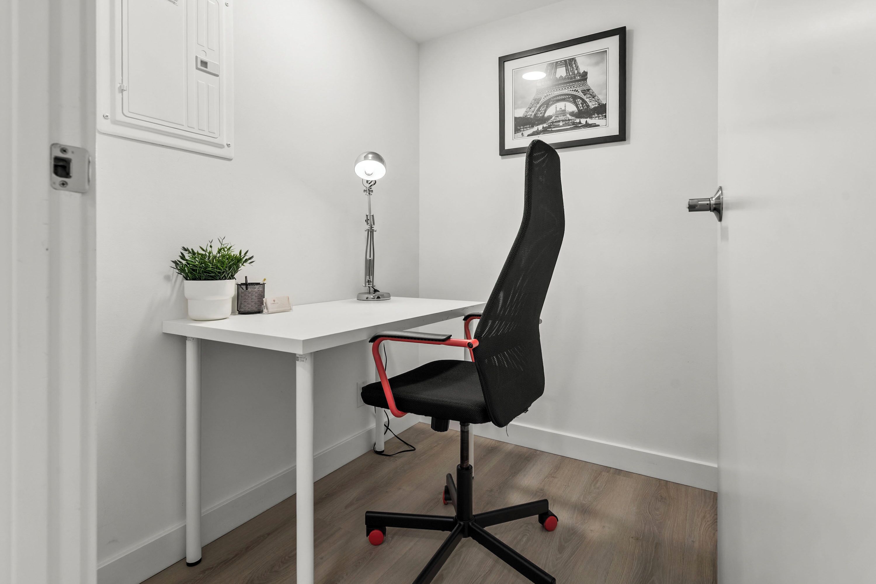 A modern office space with a white desk, a black office chair with red accents, a floor lamp, a potted plant, and a framed picture of the Eiffel Tower on the wall.