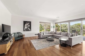 A modern living room with a sectional sofa, a coffee table, a TV, and a large window with a view of greenery. - Bush to Bay: Nature, Surf Beach & Wineries (Yallingup)