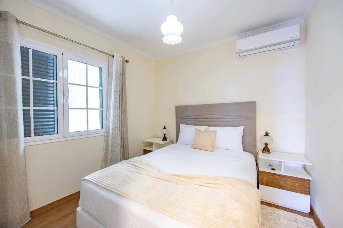 A bedroom with a large bed, bedside tables, a nightstand, an air conditioning unit, and a window with curtains.