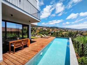 A modern outdoor pool area with wooden decking, a lounge chair, and a view of a hillside town. - CANTINHO do OUTEIRO - Aurore (Canhas)