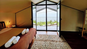 Een slaapkamer met een bed, nachtkastje, lamp en uitzicht op een balkon met planten.