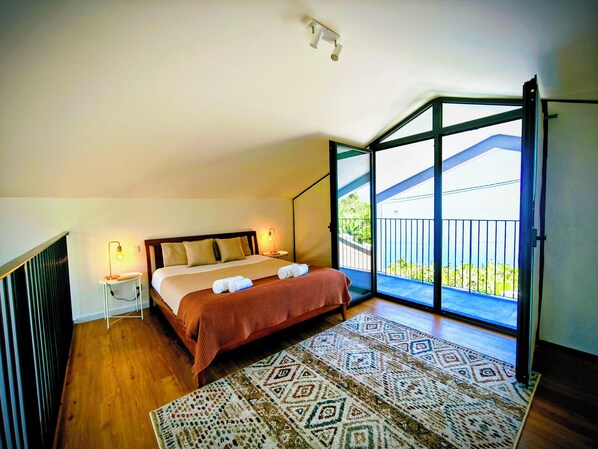 A bedroom with a large bed, two bedside lamps, a patterned rug, and a balcony with a view. - CANTINHO do OUTEIRO - Aurore (Canhas)