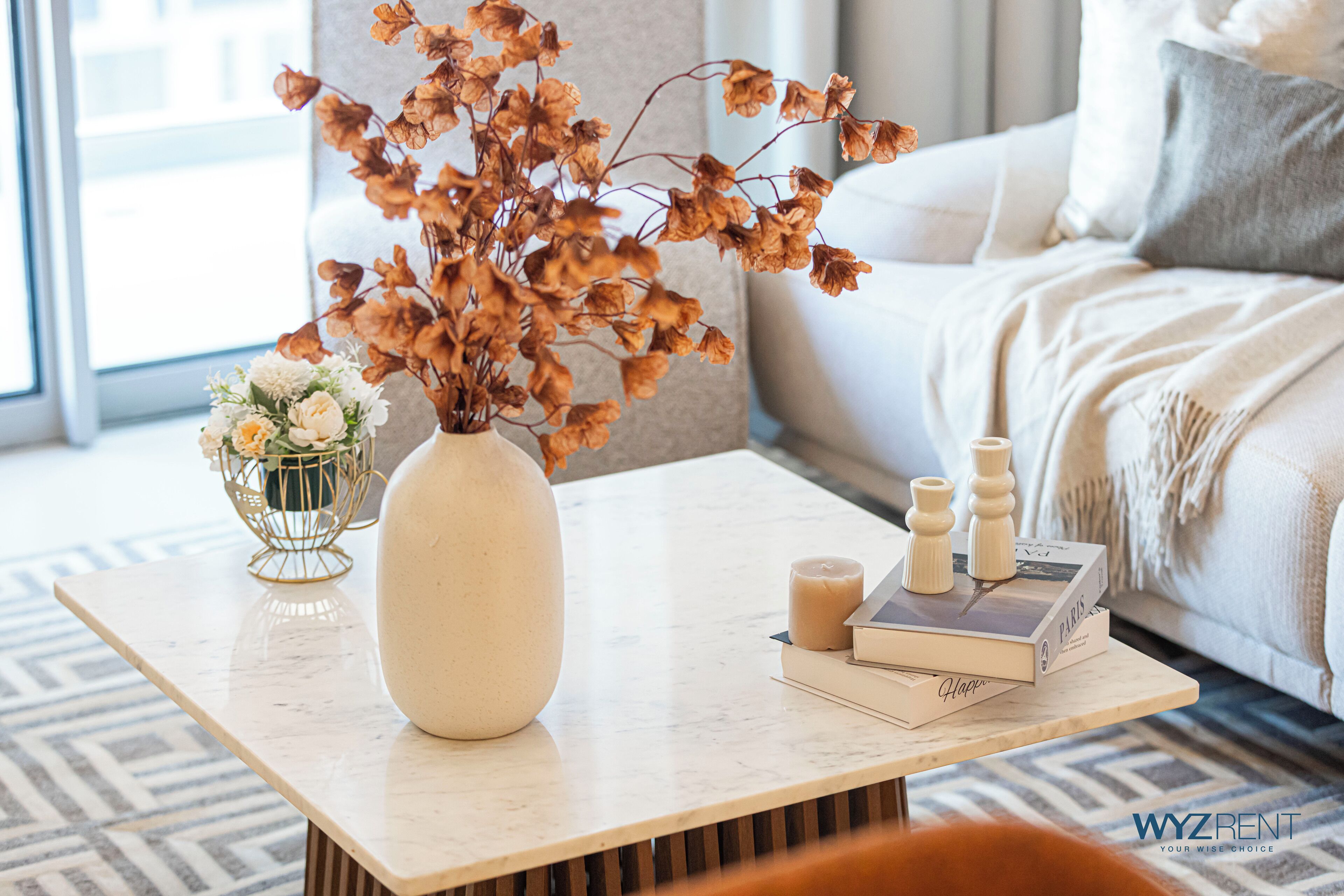 Un salon moderne avec un canapé beige, une table basse ornée d’un vase de fleurs séchées, une bougie et un livre.