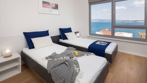 Een moderne slaapkamer met twee bedden, een groot raam met uitzicht op zee en een nachtkastje met een lamp.