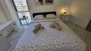 Un dormitorio con una cama, dos mesitas de noche, un ventanal con vistas a edificios y dos carteles en la pared.