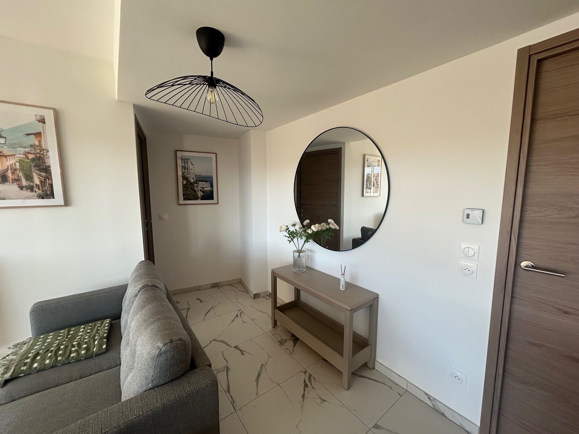 Un salon moderne avec un canapé gris, un miroir rond, une console en bois et un luminaire suspendu au plafond.
